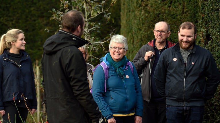 A group of people in front of a yew hedge, smiling as they listen to a guide.